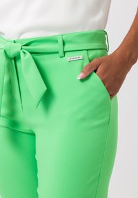 Groene, aansluitende broek van gladde stof, met een brede tailleband met strik, zijzak en een klein metalen label.