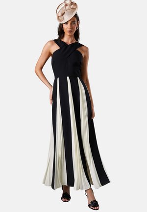 Donna che indossa un vestito maxi plissettato bianco e nero con spalline incrociate e un cappello fascinator decorativo beige.