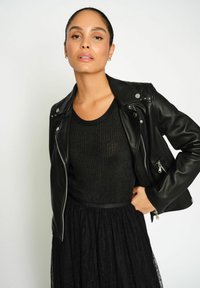 Moteris dėvi juodą odinę striukę ant juodo raukšlėto viršaus ir nėrinių sijono, pozuoja su viena ranka ant klubo prieš lygią fono.