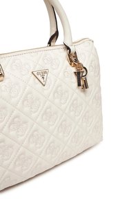 Guess laukku, joka on valmistettu vuorattusta kermavärisestä keinonahasta, kultasävyisillä metalliosilla, logoplaketilla ja koristeellisella avaimenperäsydämellä.