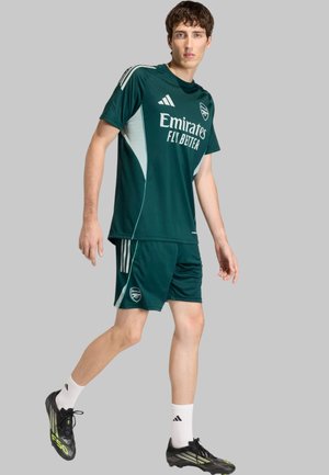 Modelo masculino caminando, vistiendo el uniforme de fútbol del Arsenal en verde oscuro con los logos de "Emirates Fly Better" y Adidas, botas negras y calcetines blancos de Adidas.