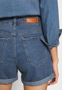Jeansshorts i medelblått med uppvikta fållar, utrustade med två bakfickor och en brun läderetikett ovanför midjan.