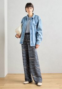 Džínová bunda, světle modrá, oversize střih; šedý top; vzorované široké kalhoty v černé barvě s modrými akcenty; drží krémovou kabelku.