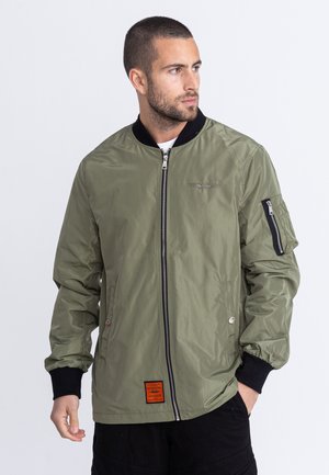 Oliwkowa bomberka z zamkiem błyskawicznym z przodu, czarnymi ściągaczami oraz zamkniętą kieszenią na lewym rękawie. Teksturowany materiał z detalami w postaci naszywki.