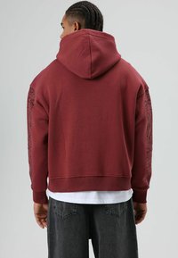 Sweat à capuche bordeaux avec une capuche, des poignets côtelés et un motif graphique le long des manches. Texture douce et coupe décontractée. Stylé avec un t-shirt blanc en dessous.