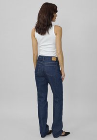 Žena so stredne dlhými hnedými vlasmi, oblečená v bielom bezrukávovom tričku, tmavomodrých rovných džínsach a čiernych balerínach, stojí pred sivým pozadím.