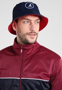 Cappello a secchi blu navy e rosso con un logo rotondo; indossato con una giacca zip maroon caratterizzata da un design a pannelli di colore e un dettaglio a striscia bianca.