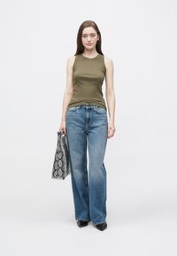 Dopasowany, bezrękawnik w oliwkowej zieleni, jasnoniebieskie szerokie spodnie jeansowe, czarne buty z ostrym noskiem, trzymająca szarą i czarną torbę w wzór skóry węża.