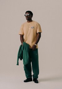 Vyras dėvintis smėlio spalvos marškinėlį, žalius sportinius kelnes, juodus batus ir akinius nuo saulės, laikantis žalią džemperį prieš pilką smėlio spalvos foną.