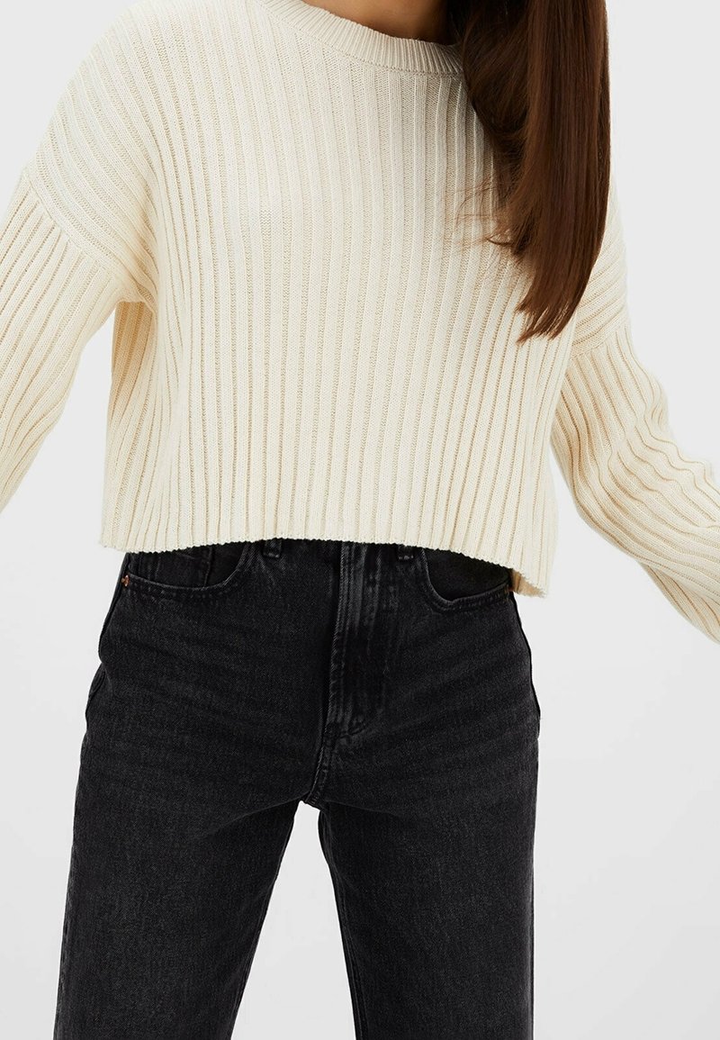 Vrouw in een crèmekleurige geribbelde cropped trui en hoogwater zwarte jeans, tegen een effen lichte achtergrond.