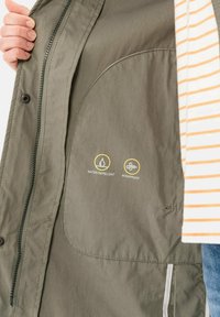 Wasserabweisende und winddichte grüne Jacke mit einem Reißverschluss, strukturiertem Gewebe und Icon-Details in der Innentasche.