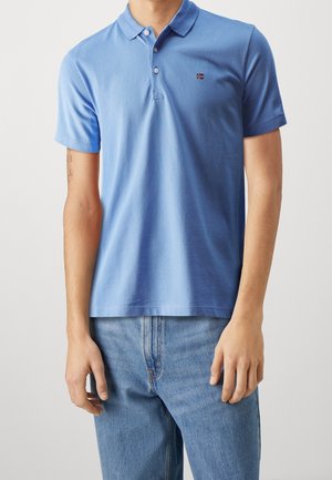 Niebieska koszulka polo z krótkim rękawem, wykonana z miękkiej bawełny, z trzema guzikami i małym logo na piersi. Zestawiona z jasnoniebieskimi dżinsami.