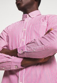 Chemise rayée rose et blanche, dotée d'un col boutonné, de manches longues avec poignets à boutons, et d'une seule poche poitrine. Tissu en coton.