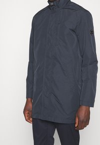 Marinblå vattentät jacka med hög krage, framknäppning och sidofickor. Har en logotyp på ärmen och en slät, matt yta.