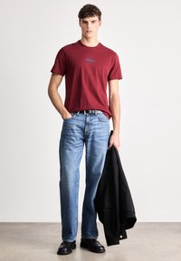 Punane lühikeste varrukatega t-särk sinise logo ja kergete siniste sirgete pükstega, mustade jalanõudega, hoiab musta jope.