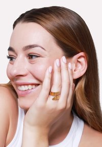 Goldfarbener Metallring mit zwei gewölbten Bändern, der an einer Hand getragen wird. Die Haut ist glatt und hell, die Nägel sind kurz mit einem hellen Nagellack, das Haar ist wellig und braun.
