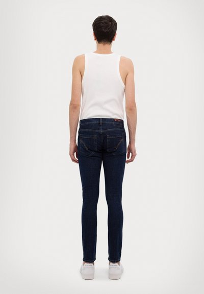 Jeans skinny blu scuro con una leggera elasticità, tasche standard, cuciture arancioni a contrasto, indossati con una canotta bianca semplice e sneakers bianche.