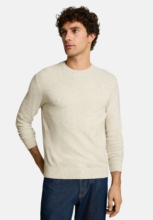 Jonge man met krullend haar die een lichtbeige gebreide trui en blauwe spijkerbroek draagt, staand met één hand achter zijn rug.