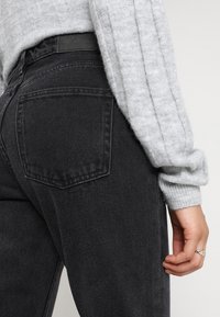 Svarta högmidjade jeans med sydda bakfickor och en läderetikett. En grå ribbad tröjärm är delvis synlig längst upp.