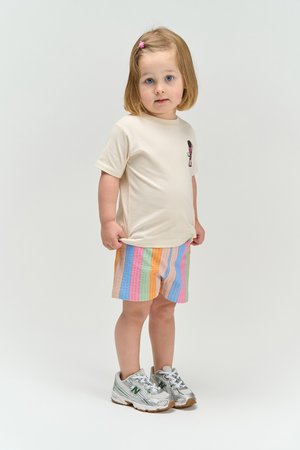 Vastsündinu roosa juuksenõelaga, kandes beežikat t-särki ja värvikirevaid triibulisi lühkse, seisab hõbedastes tossudes heledal taustal.