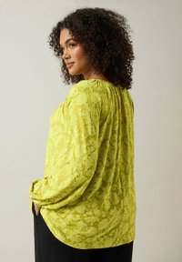 Limegrön blus med texturerat blommönster, flödande design, långa ärmar och rynkad halsringning, i kombination med svarta byxor.