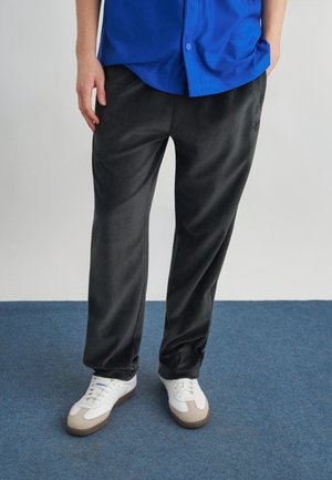 Personne portant un pantalon en tissu doux noir, des baskets blanches et une chemise bleue boutonnée, debout sur un sol recouvert de moquette bleue.