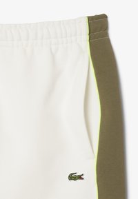Jogging blanc avec une taille élastique, panneaux latéraux vert olive et accents jaune fluo ; présente un petit logo de crocodile vert.