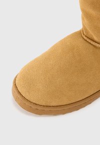 Tan ruskinds ankelstøvle med rund tå, blød tekstur og gummisål med en syet kantdetalje.