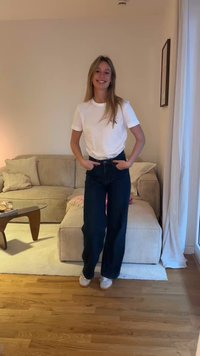 Weißes Baumwoll-T-Shirt mit Logo, hoch taillierte dunkle Denim-Hose und weiße Sneaker. Das Modell steht mit den Händen in den Taschen vor einem neutralen Hintergrund.