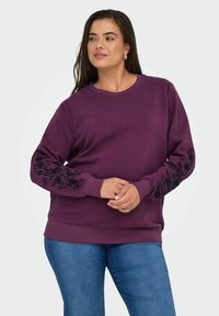 Sweatshirt violet à manches longues avec broderie florale noire au niveau des poignets. Col rond classique, tissu doux et coupe décontractée.