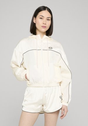 Młoda kobieta o prostych ciemnych włosach nosi kremową kurtkę z kapturem na zamek oraz dopasowane szorty z czarnymi lamówkami i logo, z rękami w kieszeniach.