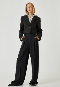 Blazer negro corto con un solo botón y una sudadera gris con cremallera debajo; combinado con pantalones anchos negros y zapatos negros.