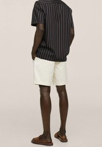 Persona in piedi con le mani nelle tasche, che indossa una camicia nera a maniche corte con sottili righe verticali, pantaloni siopi bianchi e infradito marroni.