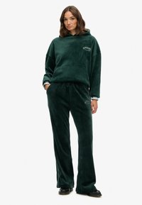 Grøn fleece træningsdragt med hættejakke og pants med snoretræk. Har et logo på brystet og en sidelæg på bukserne.