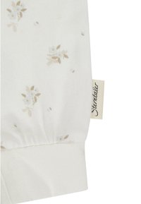 Weißer Stoff mit kleinen beigen Blumenmustern und einem angenähten cremefarbenen Etikett mit geschwungener schwarzer Schrift am umgeschlagenen Rand.