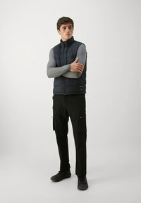 Marineblå vattert vest over en tettsittende grå langermet skjorte, kombinert med svarte cargobukser og svarte sko, stående mot en lys bakgrunn.