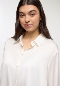 Camicia bianca con colletto, caratterizzata da una texture liscia e da una leggera lucentezza. I bottoni dorati valorizzano il design senza distogliere l'attenzione dal tessuto.