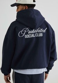 Marineblå hættetrøje med hvid broderet tekst "Prohibited SOCIAL CLUB". Bomuldsstof, afslappet pasform, diskrete ribbede manchetter og kant.