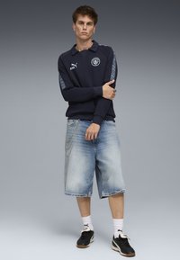 Polo a maniche lunghe blu navy con logo sul petto e strisce laterali a motivo, abbinata a pantaloncini di denim chiaro e sneakers nere, con dettagli a catena argentata.