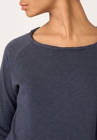 Asmuo dėvi tamsiai pilką tekstūruotą ilgomis rankovėmis marškinius su neapdorotu apykaklės kraštu, matoma kaklo ir žandikaulio dešinė pusė.