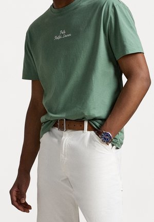 Mężczyzna ubrany w zieloną koszulkę Polo Ralph Lauren wpuszczoną w białe spodnie z brązowym paskiem i zegarkiem na nadgarstek z niebieską tarczą.