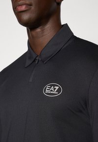 Fekete póló gombos gallérral és texturált felszíni kialakítással. Az elé az EA7 Emporio Armani logó fehér színben található a bal mellkasnál.