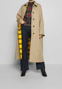 Manteau trench beige avec des boutons noirs, doublure à carreaux jaunes ; porté par-dessus un haut à carreaux multicolores et un pantalon en denim foncé. Bottines noires.