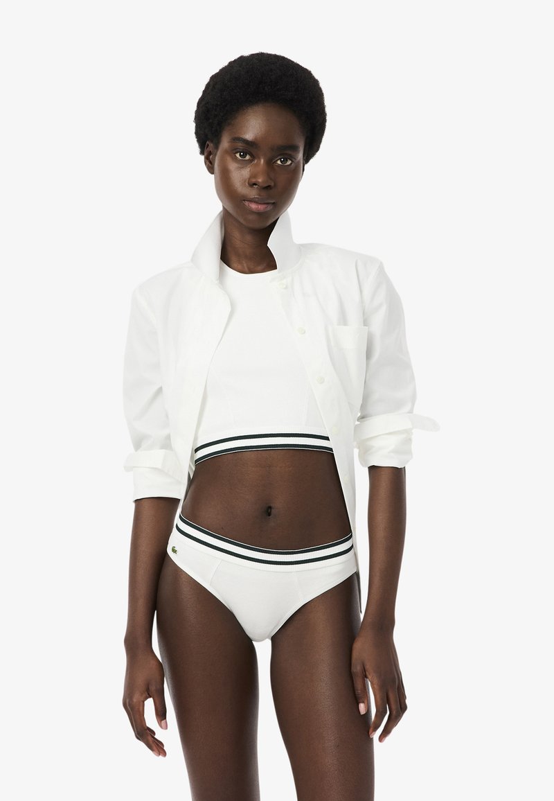 Femme noire aux cheveux courts portant un débardeur court blanc, un sous-vêtement blanc avec une ceinture rayée noir et blanc, et une chemise blanche ouverte.