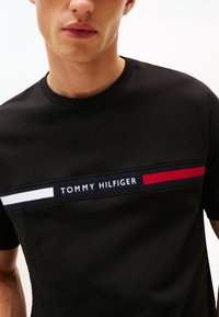 Moški, ki nosi črno majico s črnim vratom in horizontalno pasjo z logotipom Tommy Hilfiger v belem, mornarsko modrem in rdečem čez prsni koš.
