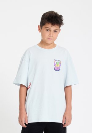 Jongen met donker haar staat vooruit kijkend, draagt een oversized lichtblauw T-shirt met een kleurrijke cartoonafbeelding op de borst en zwarte broek.
