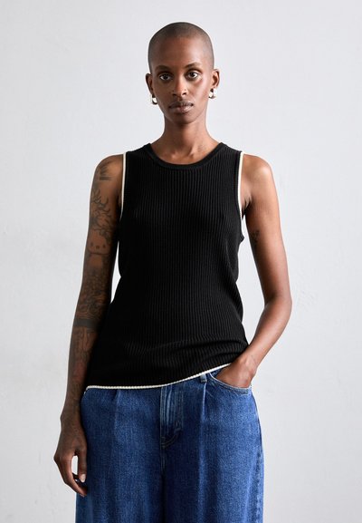 Sort ribbet tank top med hvid kant ved skuldrene, parret med løse blå denimbukser. Huden er prydet med synlige tatoveringer.