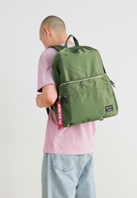 Mochila verde hecha de tela suave, con un bolsillo delantero con cremallera y una pequeña etiqueta, llevada a la espalda por una persona con una camisa rosa.
