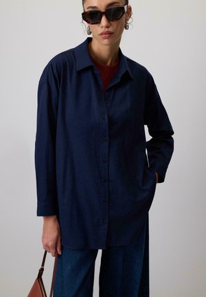 Navy blue button-up shirt with long sleeves, relaxed fit, and a collar; paired with wide-legged dark denim trousers.