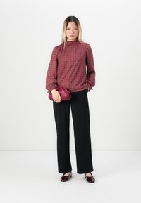 Vrouw in een gestructureerde bordeauxrode blouse, zwarte wijde broek, bordeauxrode schoenen, die een matchende bordeauxrode clutch vasthoudt tegen een witte achtergrond.